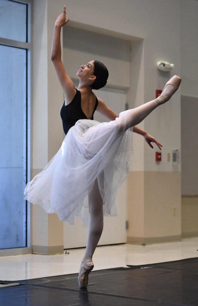 Saela Rivera-Martinez poses in attitude derriere facing away from the camera. Her arms are in an upturned fourth position. She is in a dance studio and wears a dark leotard with a white Romantic-length practice tutu