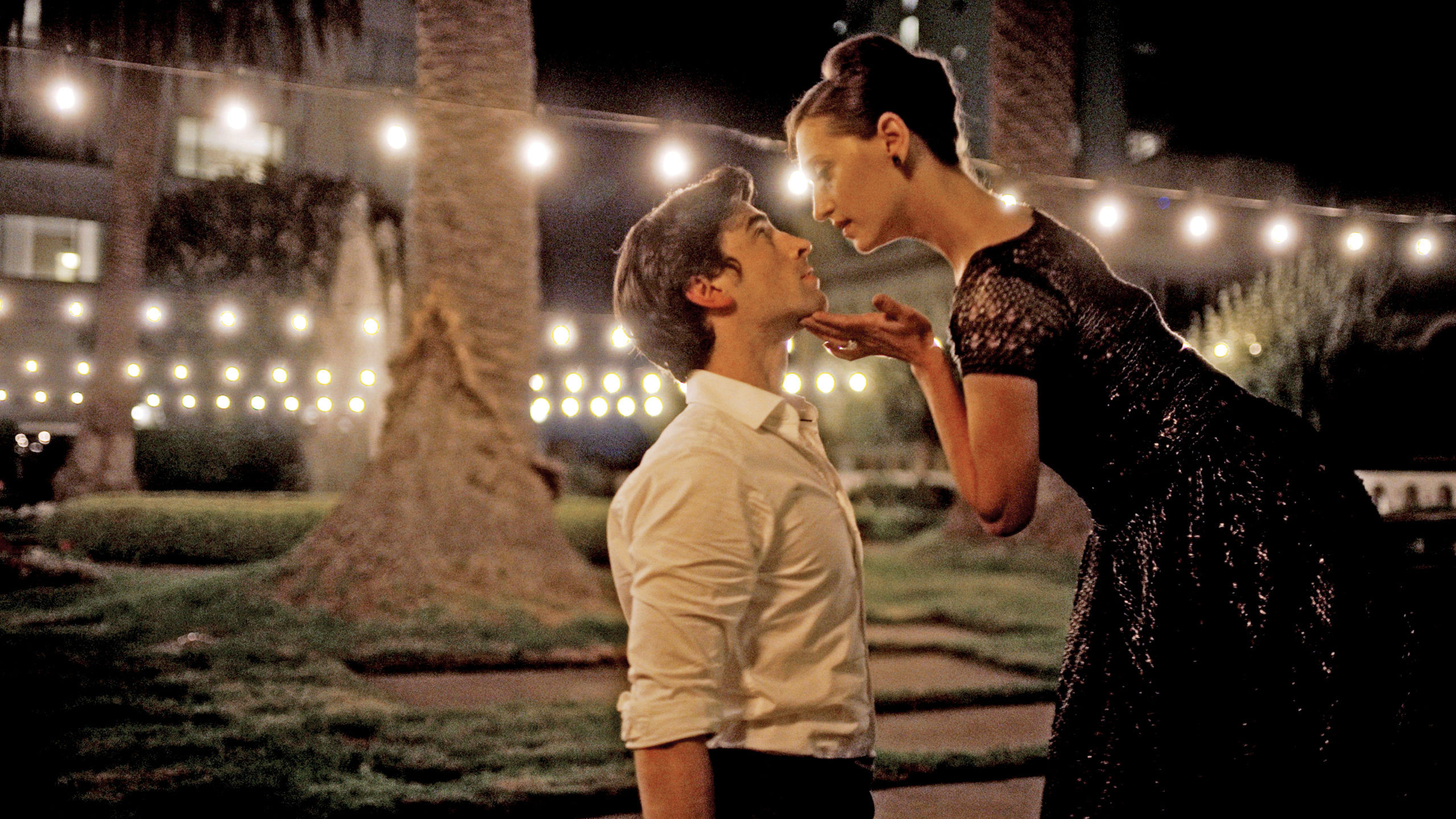 In a palm-tree lined courtyard at night, Joseph Walsh kneels onto the ground while Sarah Van Patten stands and bends towards him, lifting his chin with her left hand and looking into his eyes. Walsh wears a white button-down shirt, while Van Patten is shown in a lacy black dress, her hair in a French twist.