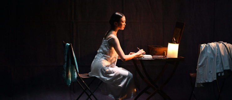 Ballerina Shiori Kase, costumed in a white nightgown, sits at a table onstage and writes a letter by candlelight. Clothes are strewn over the back of another chair close by.