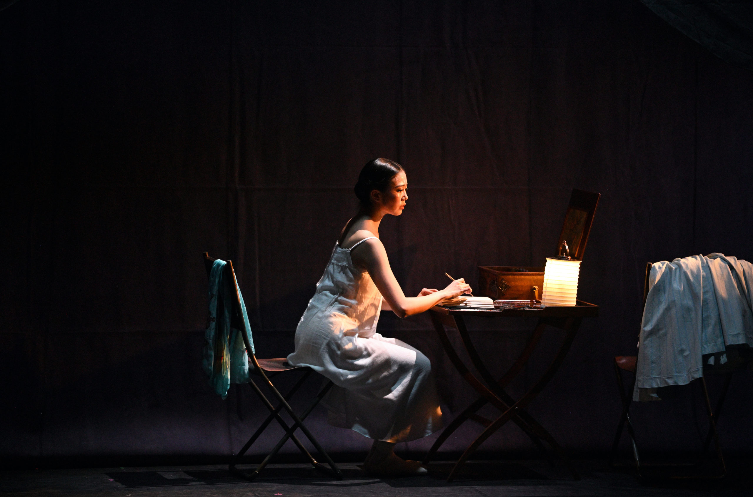 Ballerina Shiori Kase, costumed in a white nightgown, sits at a table onstage and writes a letter by candlelight. Clothes are strewn over the back of another chair close by.