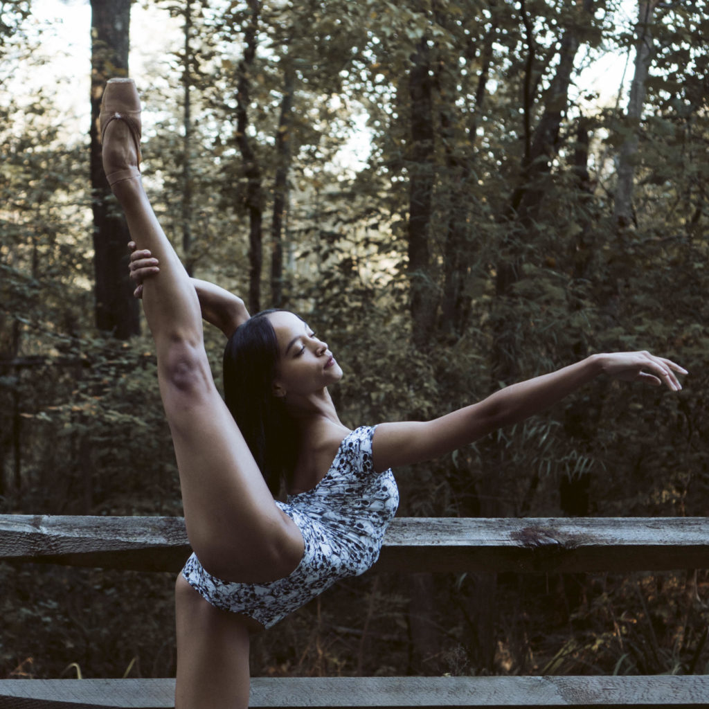 Sydney Upchurch holds her right leg in arabesque with her free left arm in front of her in arabesque. She stands in front of a fenced wooded area and wears a multi-colored leotard.
