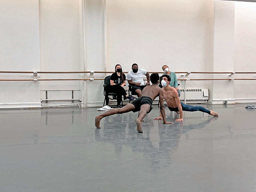 Sarah Lozoff, Christopher Rudd and Nancy Raffa, all wearing face masks, sit in chairs at the front of a large dance studio. They watch dancers Calvin Royal III and João Menegussi as they rehearse an intimate pas de deux on the floor, each propping themselves up by the hands and facing each other. The dancers are shirtless and wear face masks, with Calvin wearing dark shorts and João wearing black tights.