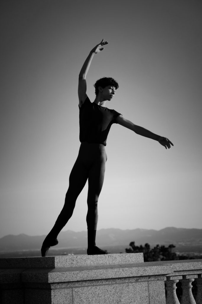 Black and white photo of Victor Galeana-Ruiz in a tendu effacé derriere. He wears dark leggings and a dark shirt. He's standing on a concrete railing in front of an outdoor background