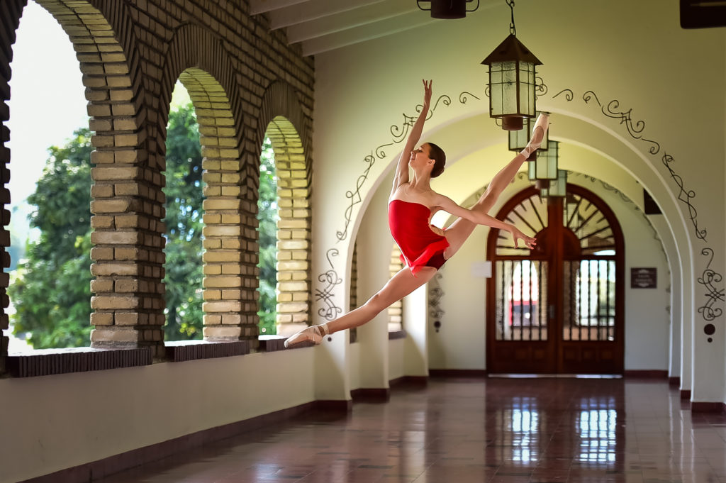 Yana Peneva jumps in a sissone en avant with her arms in a high arabesque. She's in a brightly-lit hallway that has arches into the outdoors. She wears a red leotard with a short matching skirt.