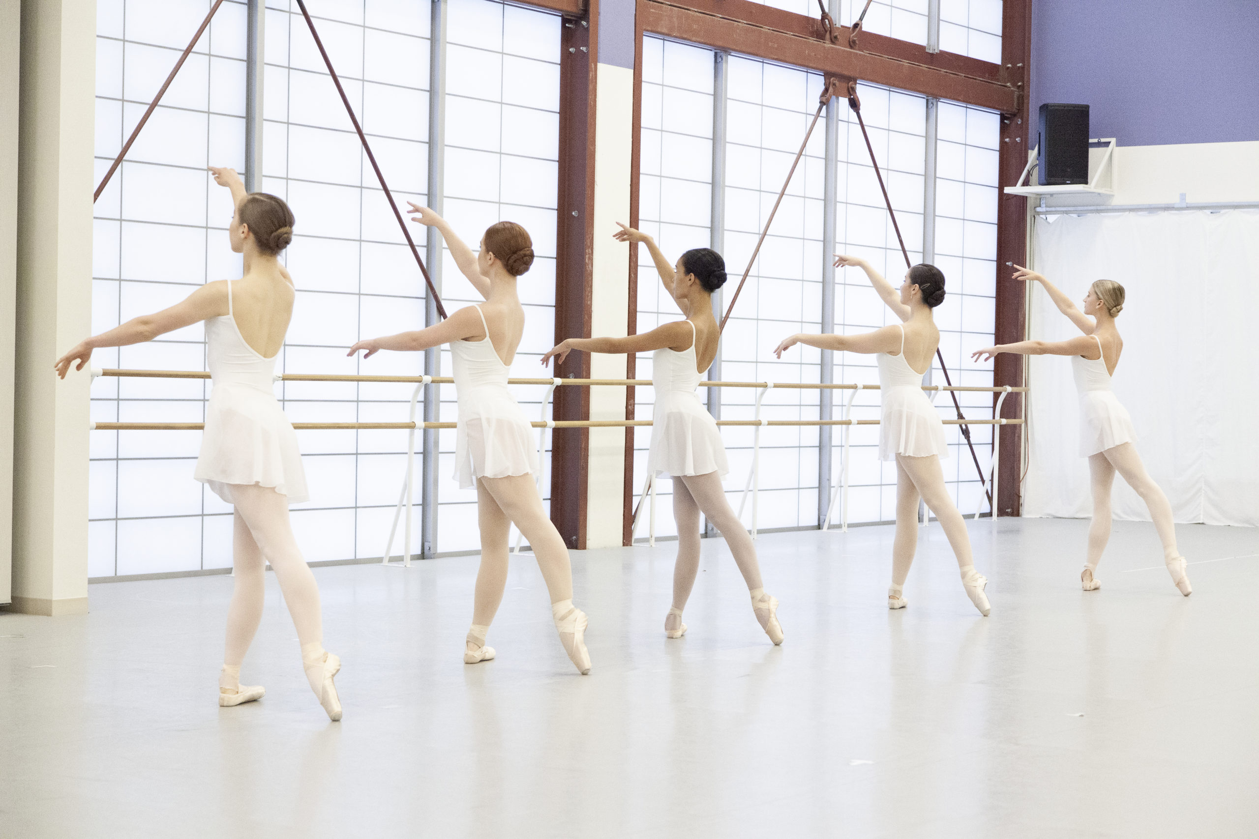 Five preprofessional female ballet dancers stand in tendu derrière facing the back of a bright studio. They are wearing white leotards and skirts and pointe shoes.