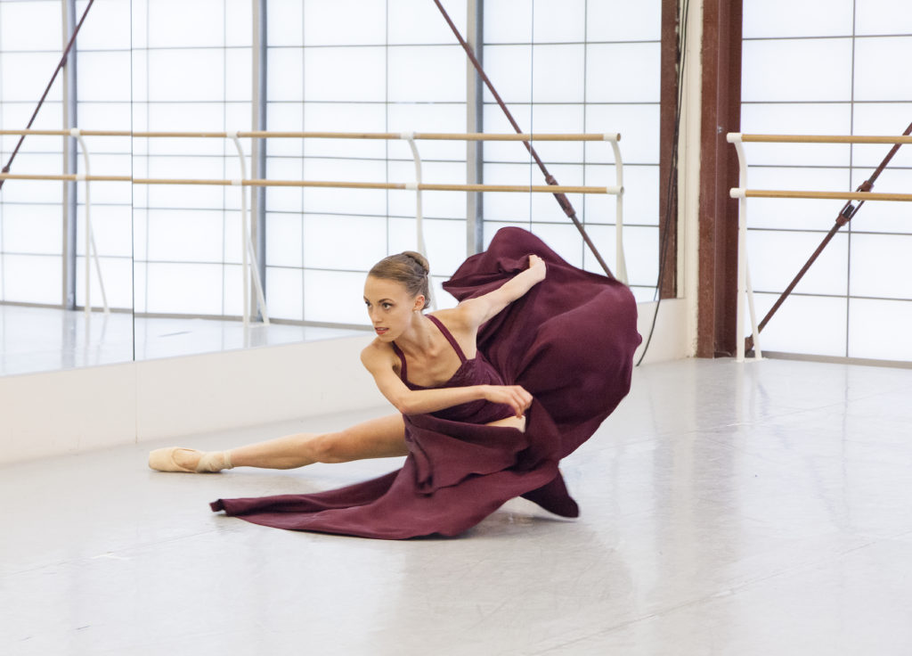 Brooke Gilliam crouches low to the ground with her right bare leg extended sideways. She is in a dance studio and wearing a long maroon dress and whipping the skirt's fabric around her torso.