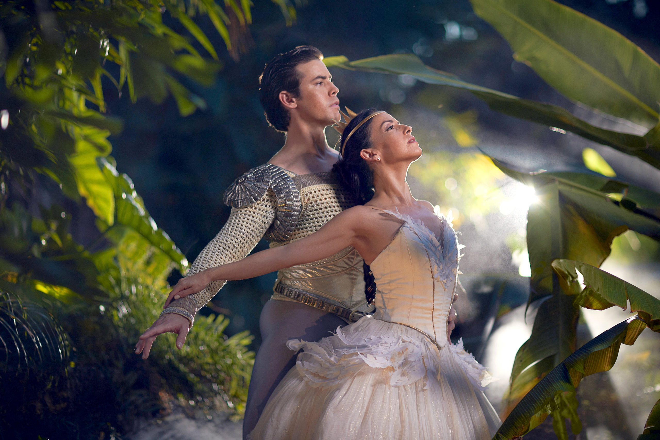 Carlos Quenedit and Katia Carranza pose in a promotional photo for the ballet "Swan Lake." Framed by lush tree branches, the pair stand in profile looking towards the right, and Carranza, wearing a white swan tutu, arches back slightly and streches her arms behind her to touch Quenedit's arms. Quenedit wears a white and silver tunic and gray tights and holds his arms out low.