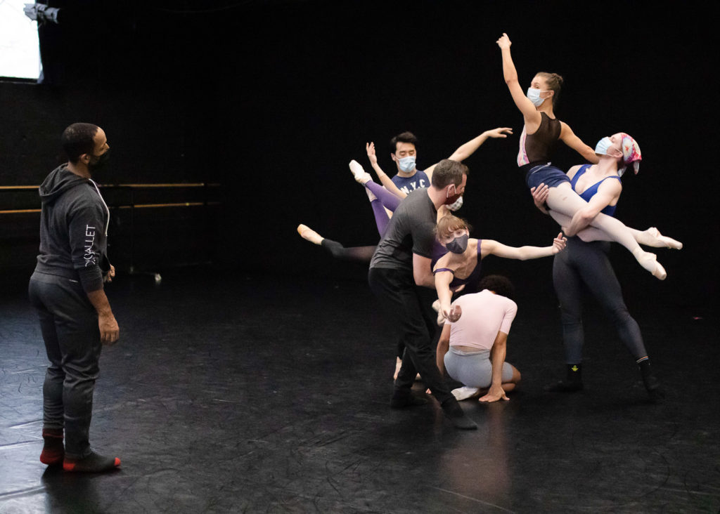In a black box rehearsal space, Ja'Malik stands in front of a group of male and female dancers as they create a tableau formation. Two men lift their female partners by the hips and legs, while a another dancer crouches low on the floor and another dances off to the side. They all wear assorted dancewear, the women in pointe shoes.