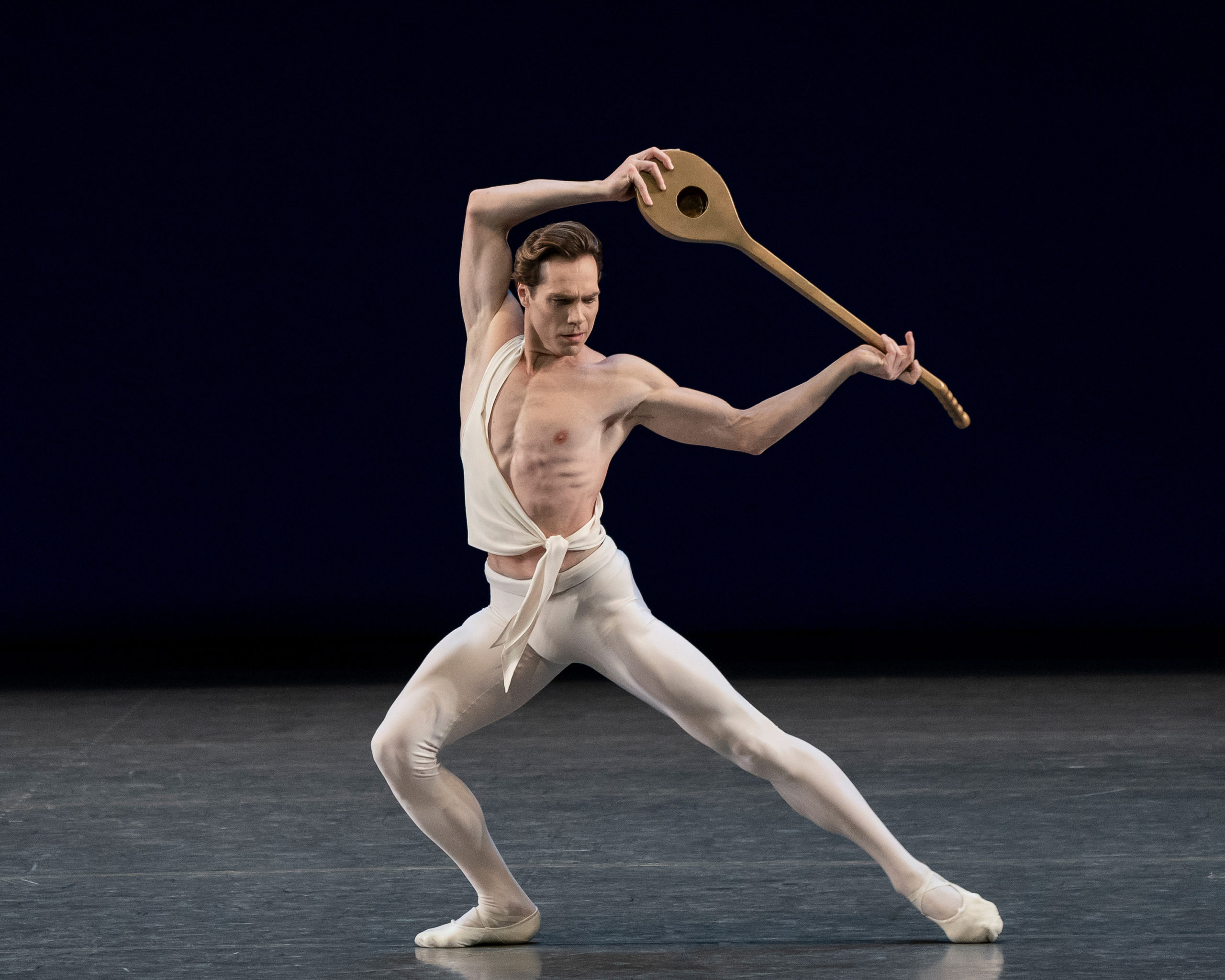 Gonzalo Garcia lunges onto his right leg onstage during a performance, stretching his left leg out to the side. He wears white tights and ballet slippers and a white toga-like shirt around his right shoulder. The left side of his chest is bare, and he lifts a lute above his head, looking down towards his left foot.