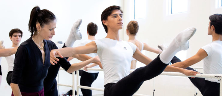 In a large, white-walled dance studio, a teenage male ballet student in a white T-shirt, black tights, and white socks and ballet slippers, does a développé devant with his right leg and holds onto a portable barre with his left hand. Next to him on his right, Arantxa Ochoa, dressed in a leotard, skirt and tights, lightly touches him on the back to correct his placement. Other dancers perform the same exercise on the other side of the barre.
