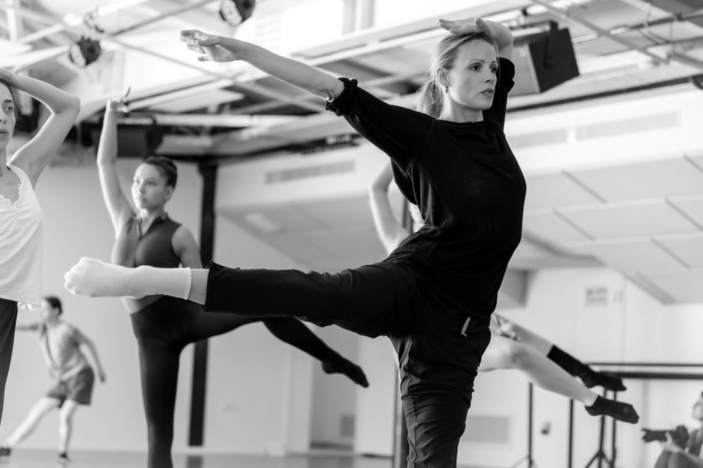 In this black and white photo, Anne Jung practices lifting her right leg to the side in a parallel side attitude. She wears a dark, long-sleeved T-shirt, dark workout pants and white socks. Behind her, other dancers practice the same movement in a large dance studio.