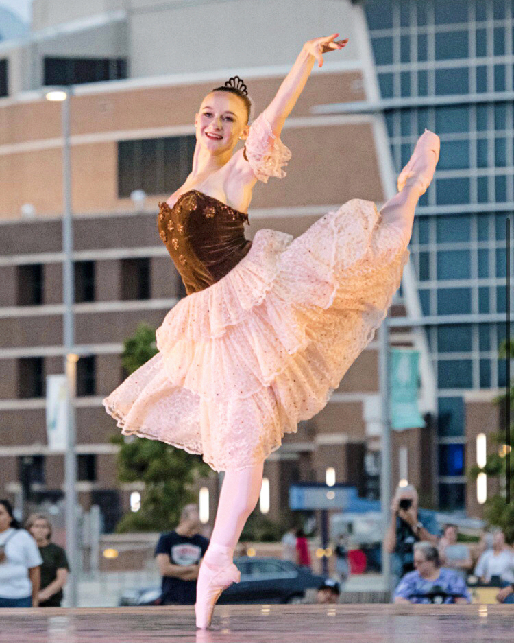 Darcey Lynn performs on an outdoor stage at twilight. She wears a lacey, knee-length tutu with a brown velvet bodice, and performs kicks her left leg behind her in a high piqué attitude, lifting her left arm high.