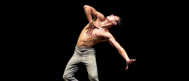 Jeffrey Cirio is shown onstage during a performance of the ballet "Creature." He is shirtless and wears gray pants and ballet shoes. Standing, he arches his body and his his head towards the right, looking at something above him. He grabds his chest with his right hand and stretches his left arm long with a flexed hand, and opens his mouth wide.