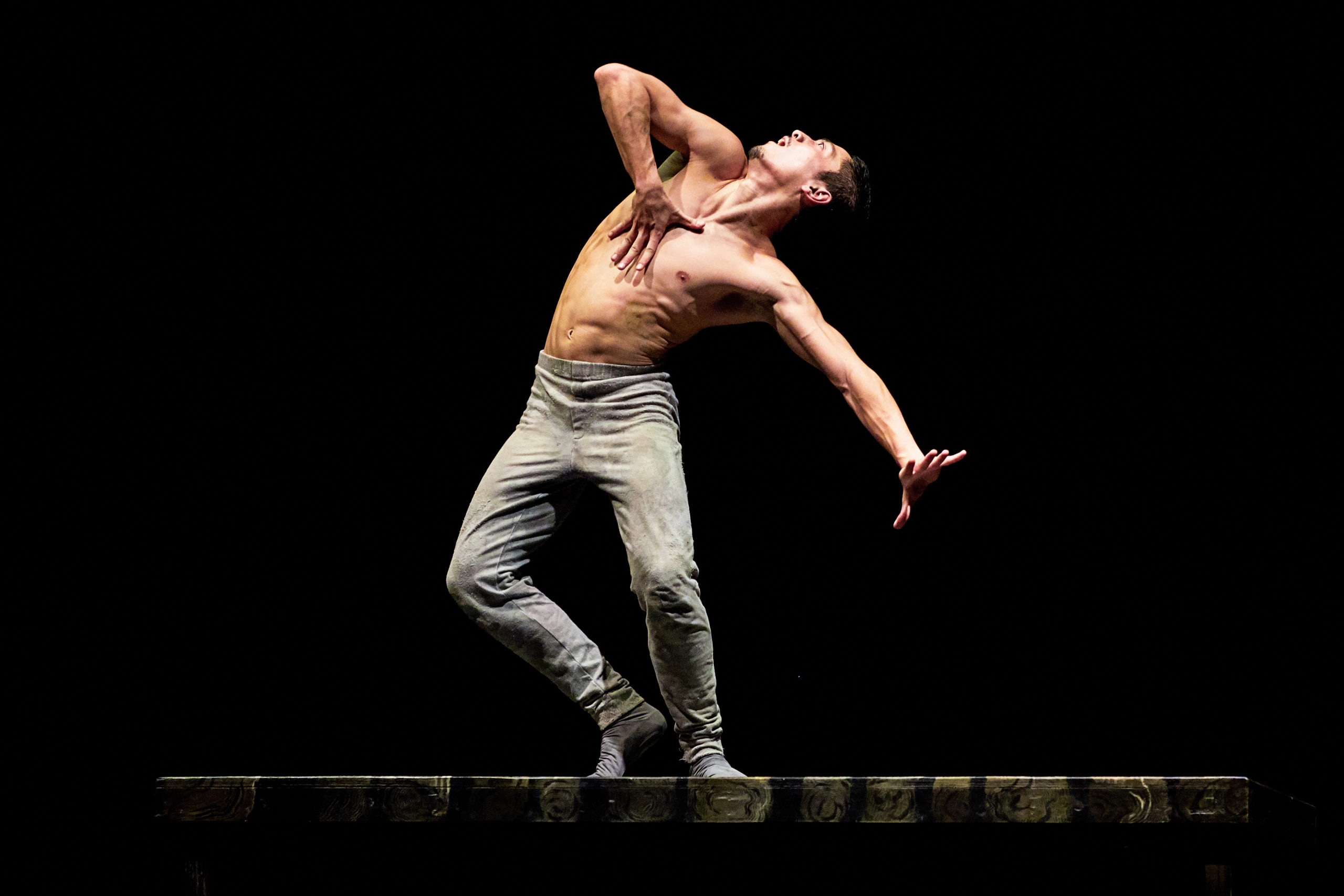 Jeffrey Cirio is shown onstage during a performance of the ballet "Creature." He is shirtless and wears gray pants and ballet shoes. Standing, he arches his body and his his head towards the right, looking at something above him. He grabds his chest with his right hand and stretches his left arm long with a flexed hand, and opens his mouth wide.