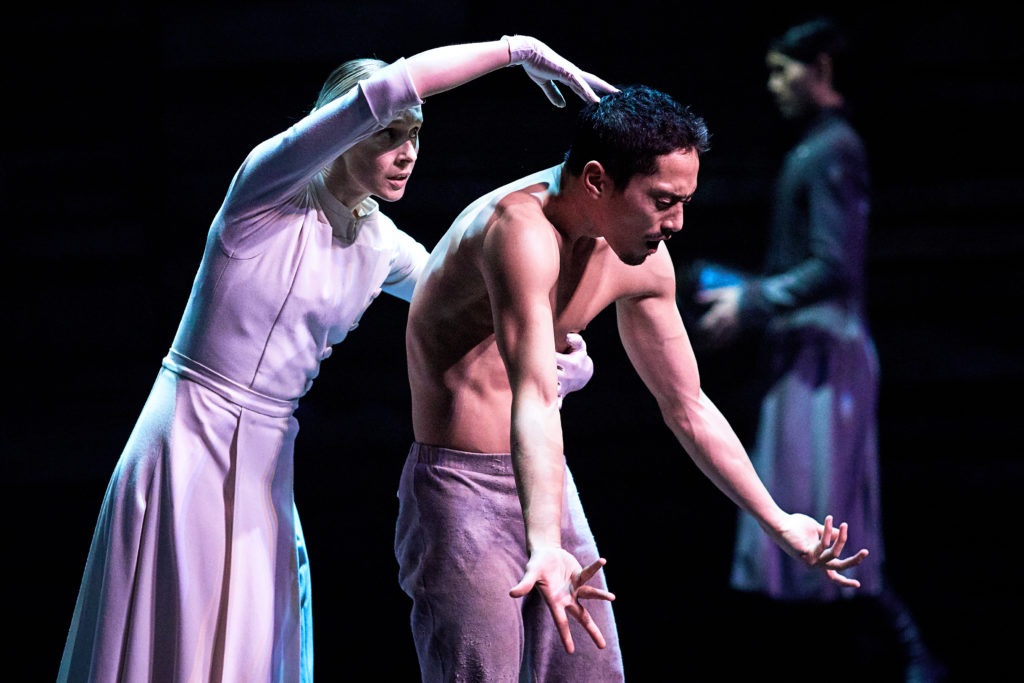 During a performance, Stina Quagebeur stands behind Jeffrey Cirio, wraps her left arm around his chest and touches the top of his head as he contracts in agony. Quagebeur is dressed in a white dress and skullcap, while Cirio is bare chested and wears gray pants.