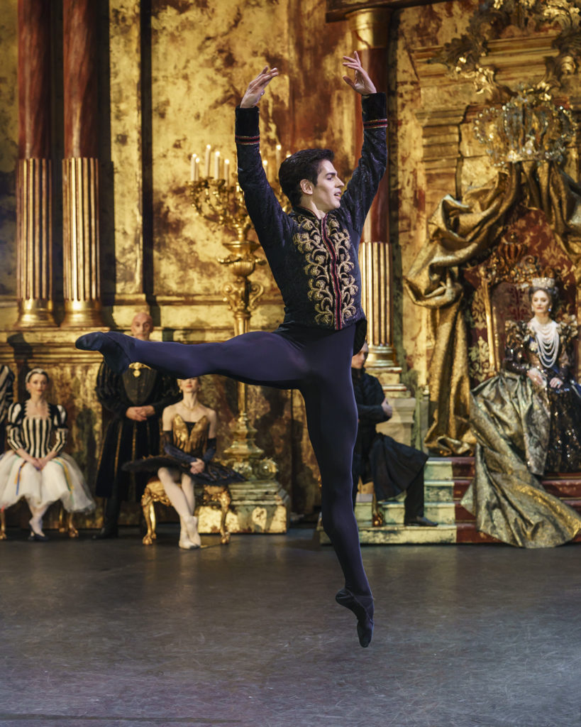 During an onstage performance of Swan Lake, Federico Bonelli does an a la seconde jump with his right leg lifted and his arms in high fifth position. He wears black tights and ballet slippers and a black tunic with gold trim. The sets behind him are grand gold pillars with candelabras and a gilded throne.