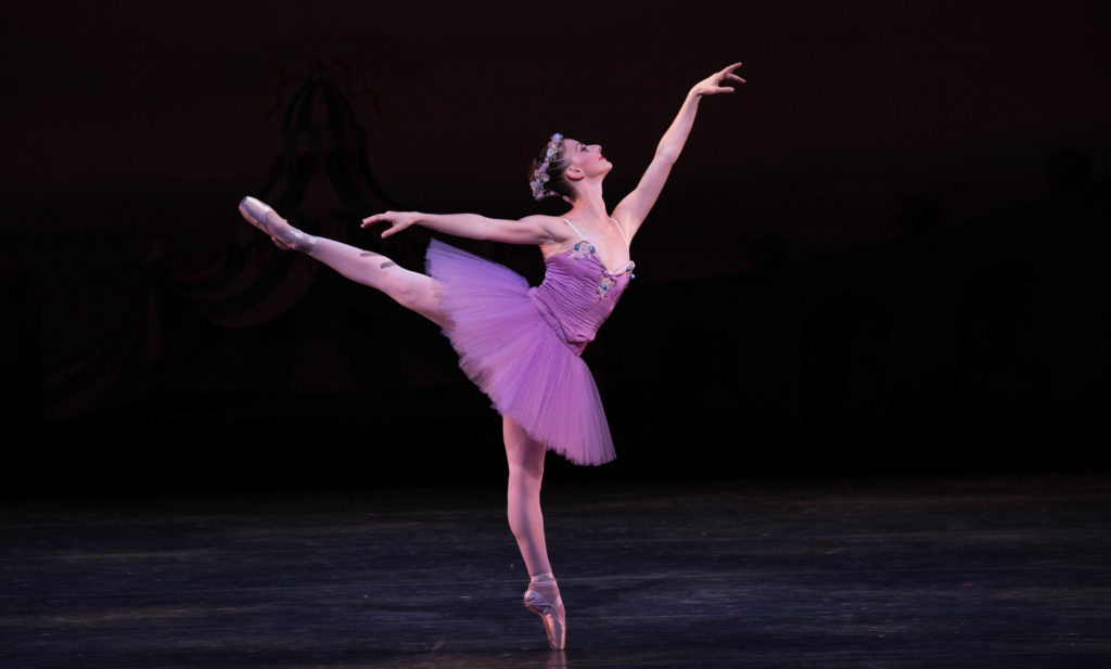 Samatha Hope Galler, wearing a purple tutu and floral-wreath headpiece, does a first arabesque on pointe during an onstage performance.