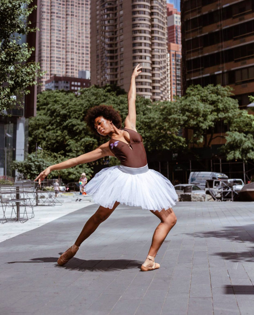 Gabriella Allyne, in a brown leotard, white practice tutu and brown pointe shoes, poses outside in a city square, surrounded by large buildings. She poses in a tendu derriere in effacé, with her right leg behind her and her left leg in plié. She holds her arms in first arabesque and arches her upper body back towards her back leg.