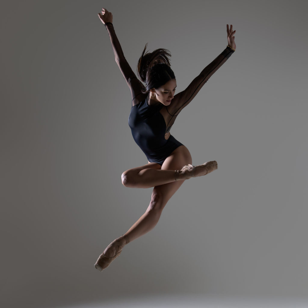 Francesca Dugarte-Jordan is captured middair in a jump with her arms spread in a high V, her left leg pointed forward and her right leg hooked over her left thigh. She is wearing a leotard and pointe shoes with bare legs.