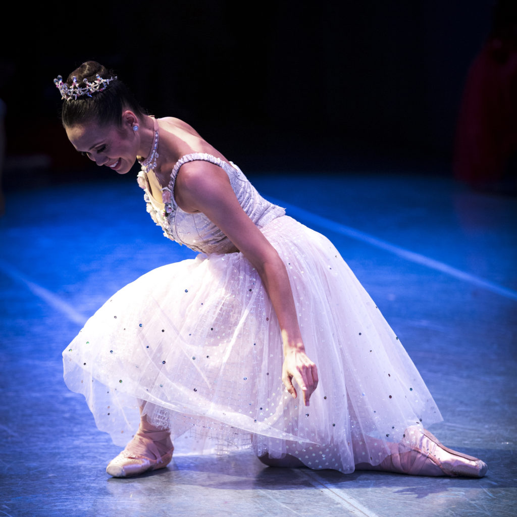 Noelani Pantastico is shown from the side taking a deep curtsey onstage. She wears a long white, glittery tutu and tiara, pink tights and pink pointe shoes, and she curtsies with her right foot behind her.