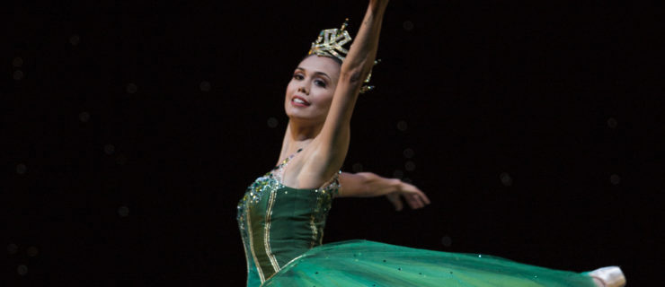 Noelani Pantastico wears an emerald green romantic-length tutu and jeweled crown during a performance. She is shown doing a piqué attitude en pointe with her left leg up behind her and her left arm raised, and she looks toward the audience with a smile. Her tutu billows around her in reaction to the turn.