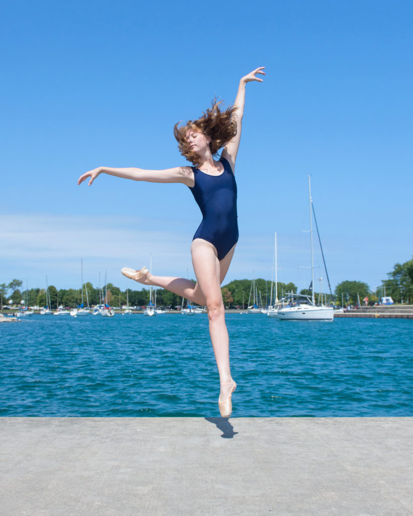 Kayla Schmitt, a teenage ballet dancer, dances outside at a marina, with water and sailboats behind her. She wears a blue leotard and pointe shoes. She does a small jump with her left leg back in a low attitude and her left arm lifted high and her right arm out to the side.