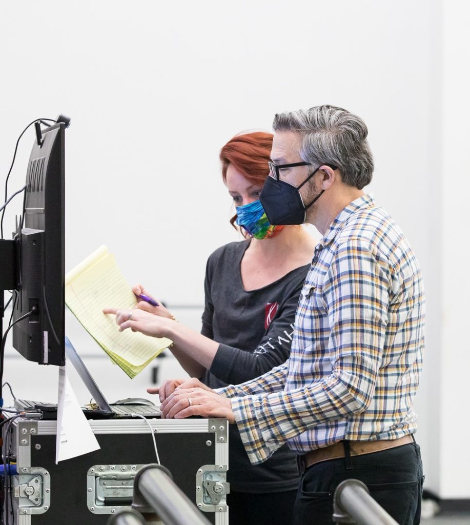 Paul Vasterling, wearing a plaid shirt and black facemask, and a ballet mistress wearing a rainbow face mask and black shirt, stand in front of a large TV monitor and review notes on a yellow pad of paper.
