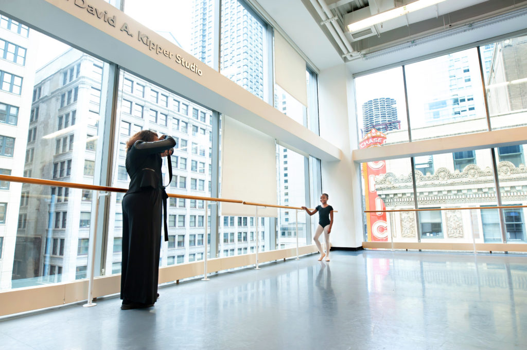 In a large dance studio with floor to ceiling windows, TaKiyah Wallace McMillian, wearing a black shirt and pants, holds her camera up to her face and takes a picture of a young female ballet student. The child, wearing a black leotard, pink tights and pink ballet slippers, holds the ballet barre with her right hand and smiles at Wallace.