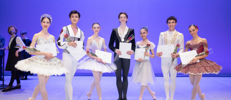 The Prix de Lausanne prize winners stand onstage in a line after the awards ceremony. There are two men and five women. The men stand in first position, the women in prep. All seven hold their certificates and smile at the camera.