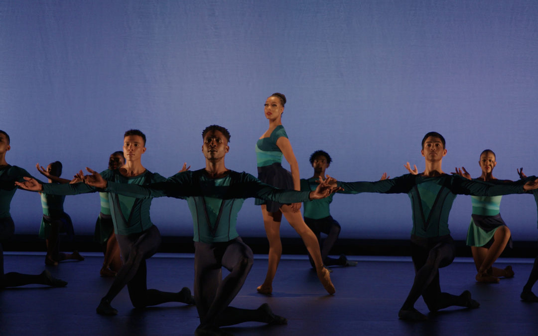 During a performance, a group of dancers form a circle onstage and kneel on their right leg with their arms outstretched to the side. In the middle, a female dancer stands in profile facing stage right with her left leg in tendu derriere and her arms low. She looks out towards the audience. All the dancers wear turqoise and blue costumes, the men in tights, tunics and ballet slippers and the women in short dance dresses and brown pointe shoes.