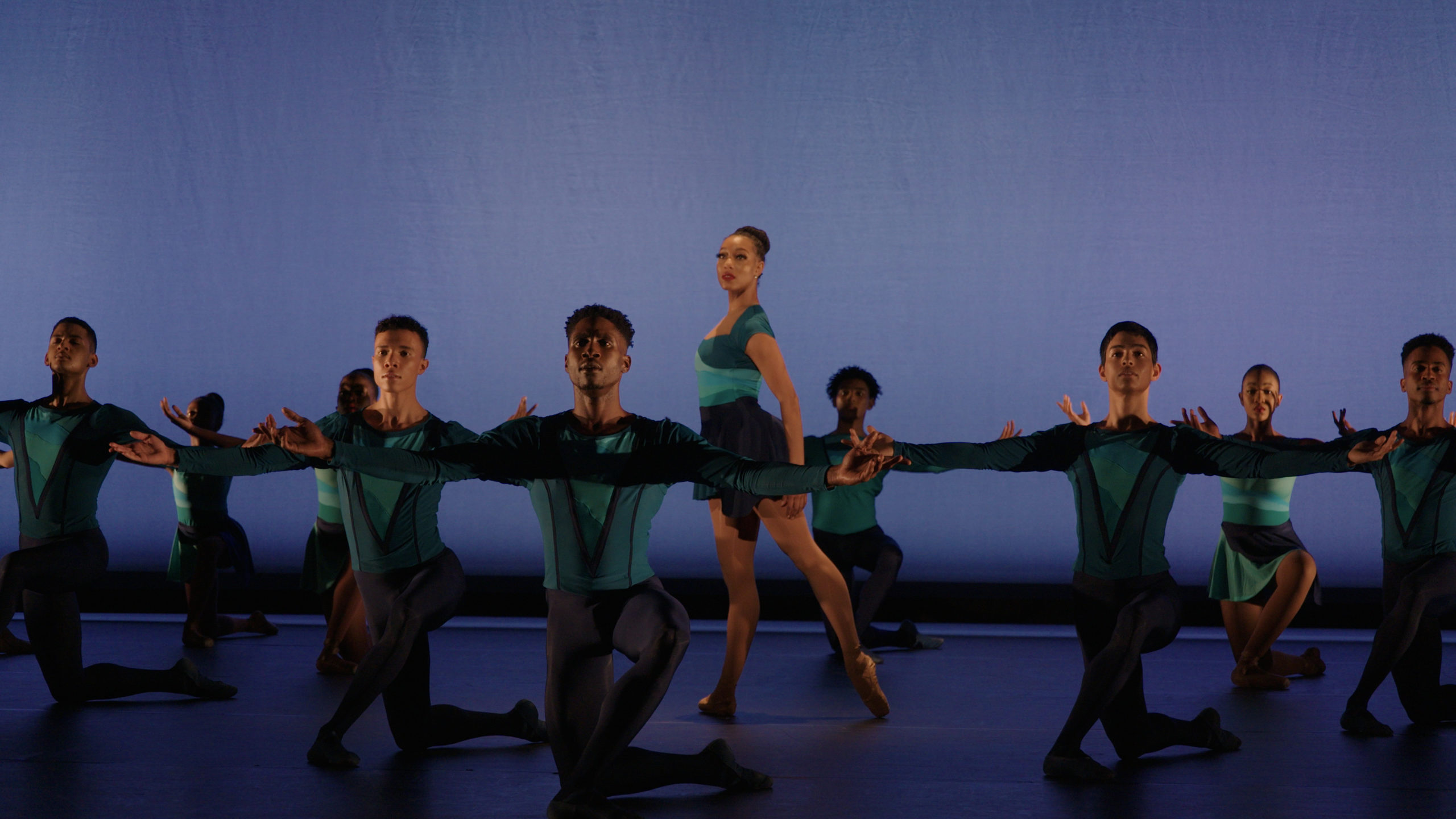 During a performance, a group of dancers form a circle onstage and kneel on their right leg with their arms outstretched to the side. In the middle, a female dancer stands in profile facing stage right with her left leg in tendu derriere and her arms low. She looks out towards the audience. All the dancers wear turqoise and blue costumes, the men in tights, tunics and ballet slippers and the women in short dance dresses and brown pointe shoes.