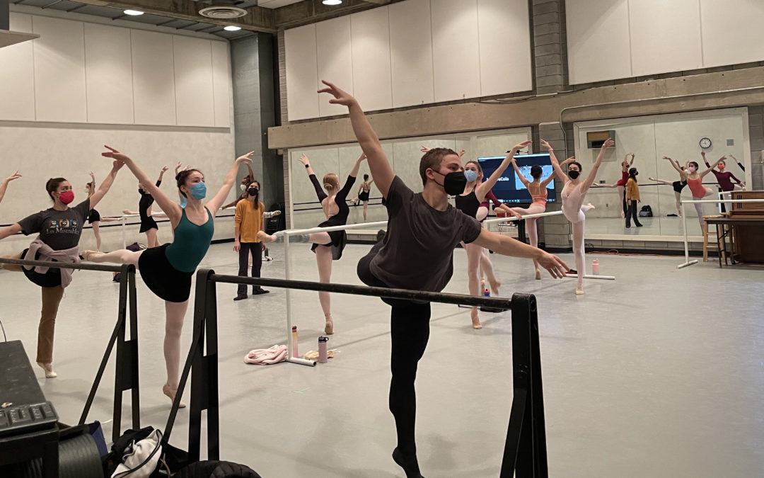 In a large, windowless dance studio, a class of male and female college-level ballet students practice balancing in arabesque at the barre. Sarah Wroth, their instructor, stands among them towards the center of the photo, and watches, She wears a yellow long-sleeved shirt and black yoga pants.