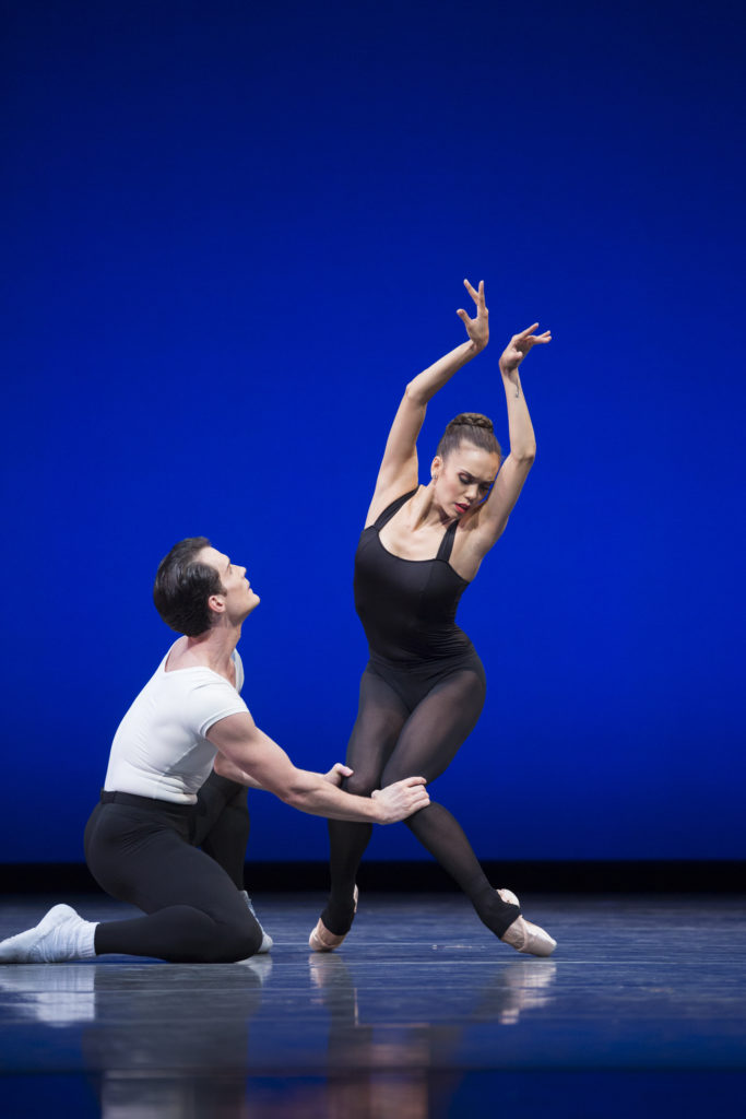 Seth Orza, wearing a white T-shirt and black tights and white sockas and ballet shoes, kneels on the ground and holds Noelani Pantastico's knees together as she balances on pointe. Pantastico pliés her knees deeply, holding them tightly together while her feet are in a wider stance, and lifts her wrists above her head, touching them together. She wears a black leotard and stirrup tights and looks down towards her left foot.