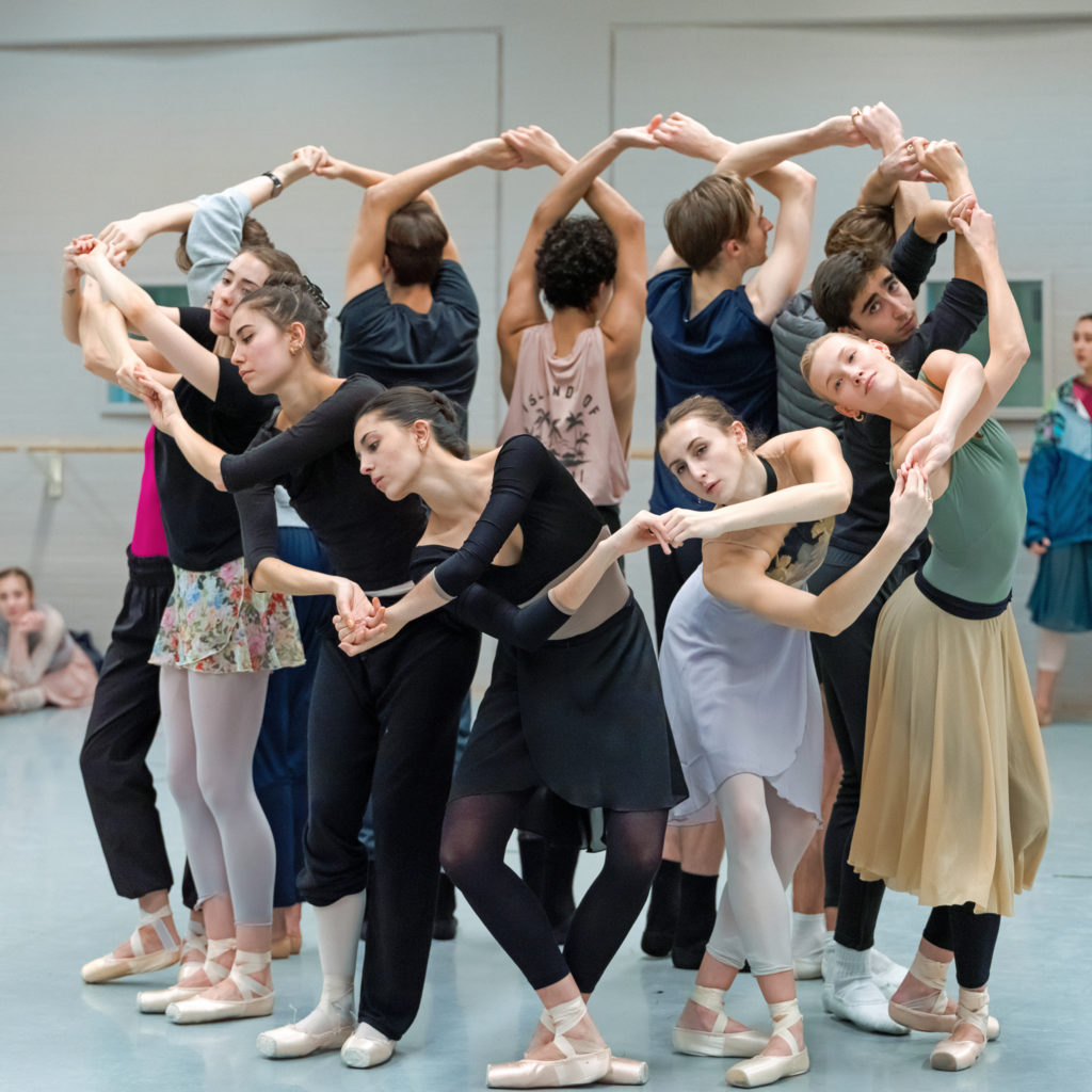 A group of 12 dancers—6 men and 6 women, form a tight circle and joined their crossed hands. The women, who form the front of the circle, bend down low while the men, facing the back of the room, keep their arms lifted. They all wear various dance practice clothes, and the women are in pointe shoes.
