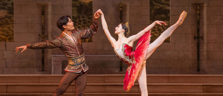 Young Gyu Choi, wearing a brown, gold and red tunic shirt, brown tights and black boots, stands on his left leg with his right extended to the side and holds Maia Makhateli's right hand as she balances in a high first arabesque on pointe. Makhateli wears a rich red tutu, gold crown and pink tights and pointe shoes. They perform onstage in front of a shallow stone staircase.
