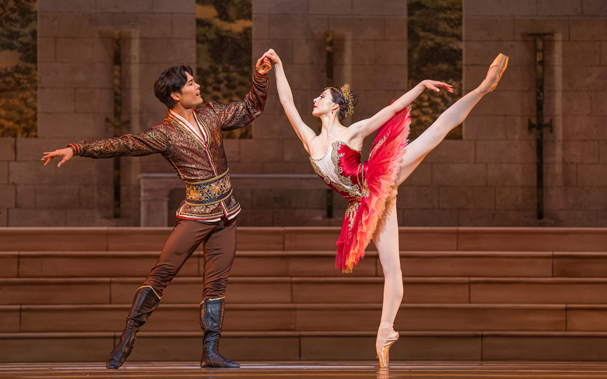 Young Gyu Choi, wearing a brown, gold and red tunic shirt, brown tights and black boots, stands on his left leg with his right extended to the side and holds Maia Makhateli's right hand as she balances in a high first arabesque on pointe. Makhateli wears a rich red tutu, gold crown and pink tights and pointe shoes. They perform onstage in front of a shallow stone staircase.