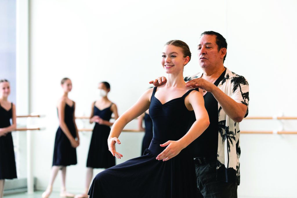 A teenage female dance student is shown from the thigh up with her arms in first position and her right leg lifted in retiré. She wears a black leotard and long black skrit. Behind her, Claudio Muñoz lighty adjusts her shoulders with his hands. He wears a white Hawaiian shirt with a black palm tree pattern, a black V-neck T-shirt and black pants.