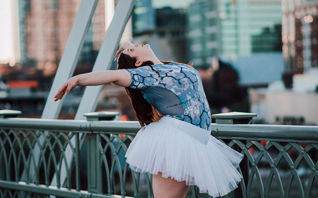 Colleen Werner stands outside on a city bridge and wears a blue leotard with a black and white pattern and a white practice tutu. She stands in paralelle on pointe (not shown) and bends backward at the waist looking up at the sky, with her arms in a V position. Her long brown hair falls down her back.