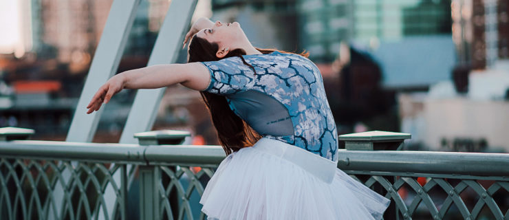 Colleen Werner stands outside on a city bridge and wears a blue leotard with a black and white pattern and a white practice tutu. She stands in paralelle on pointe (not shown) and bends backward at the waist looking up at the sky, with her arms in a V position. Her long brown hair falls down her back.
