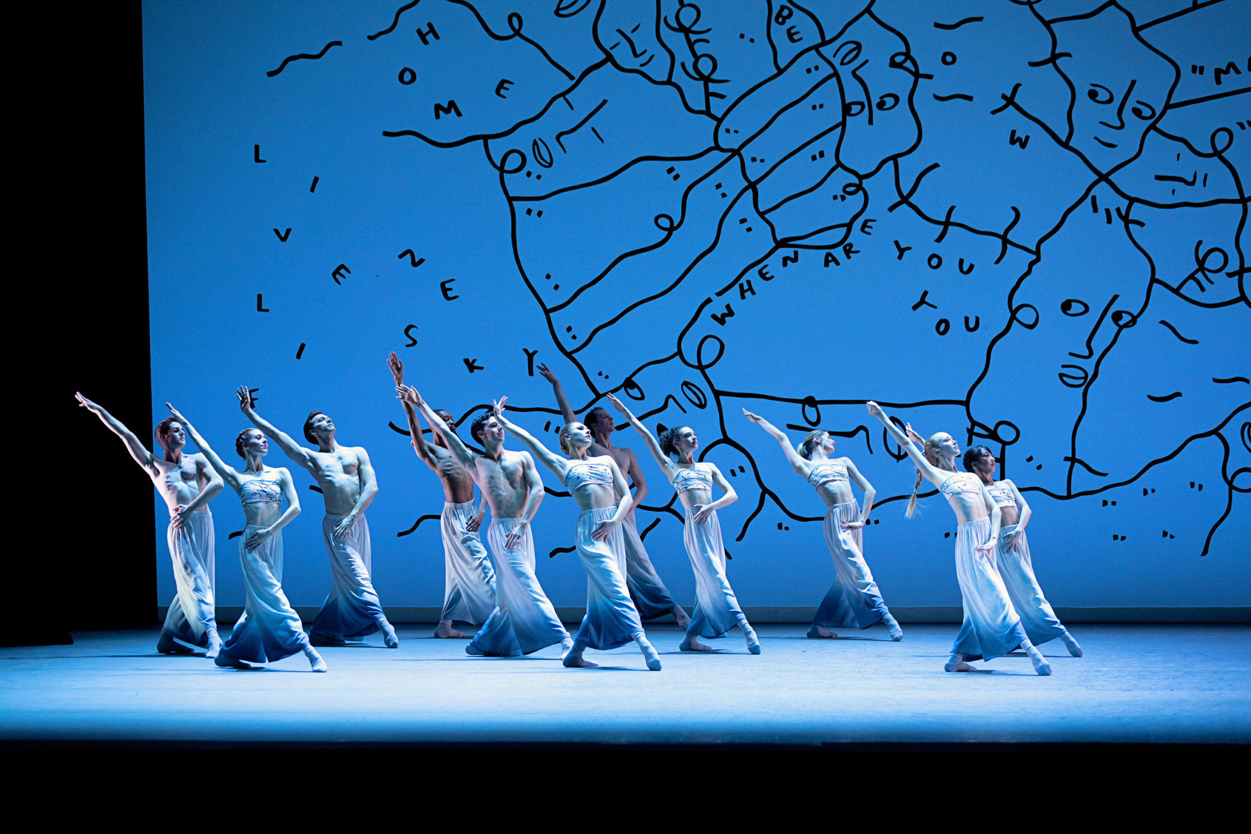 During a performance onstage, a group of 11 dancers pose in a dramatic plié tendu devant with their right leg extended and their right arm reaching high above their head and slightly behind them. Their left hand is crossed over to touch their right hip. The dancers wear long white and blue ombre skirts, and the women wear matching bandeaus. Behind them is a bright blue backdrop with a modern art design.