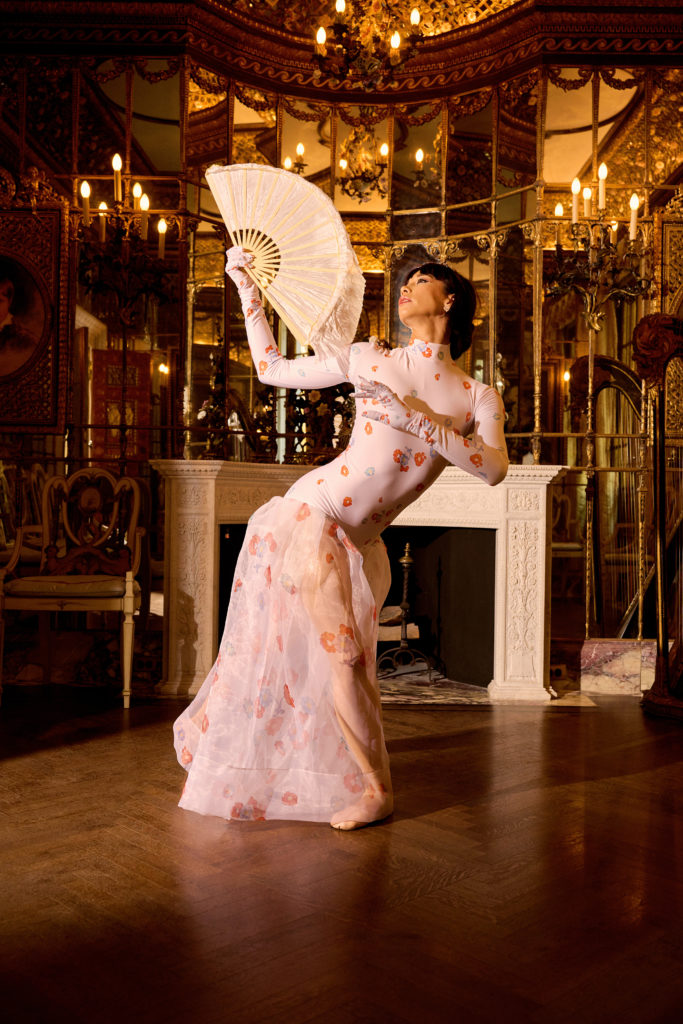In a gilded, mirrored sitting room lit by candelabras, Georginan Pazcoguin stands on her right leg in plié, extending her left leg behind her in tendu croisé, and twists her upper body dramatically to the right. She holds a large fan in her right hand and looks up at it. She wears a light-colored costume with an orange flower pattern; the top is a long-sleeved turtle neck leotard and gloves, with a sheer floor-length skirt attached.
