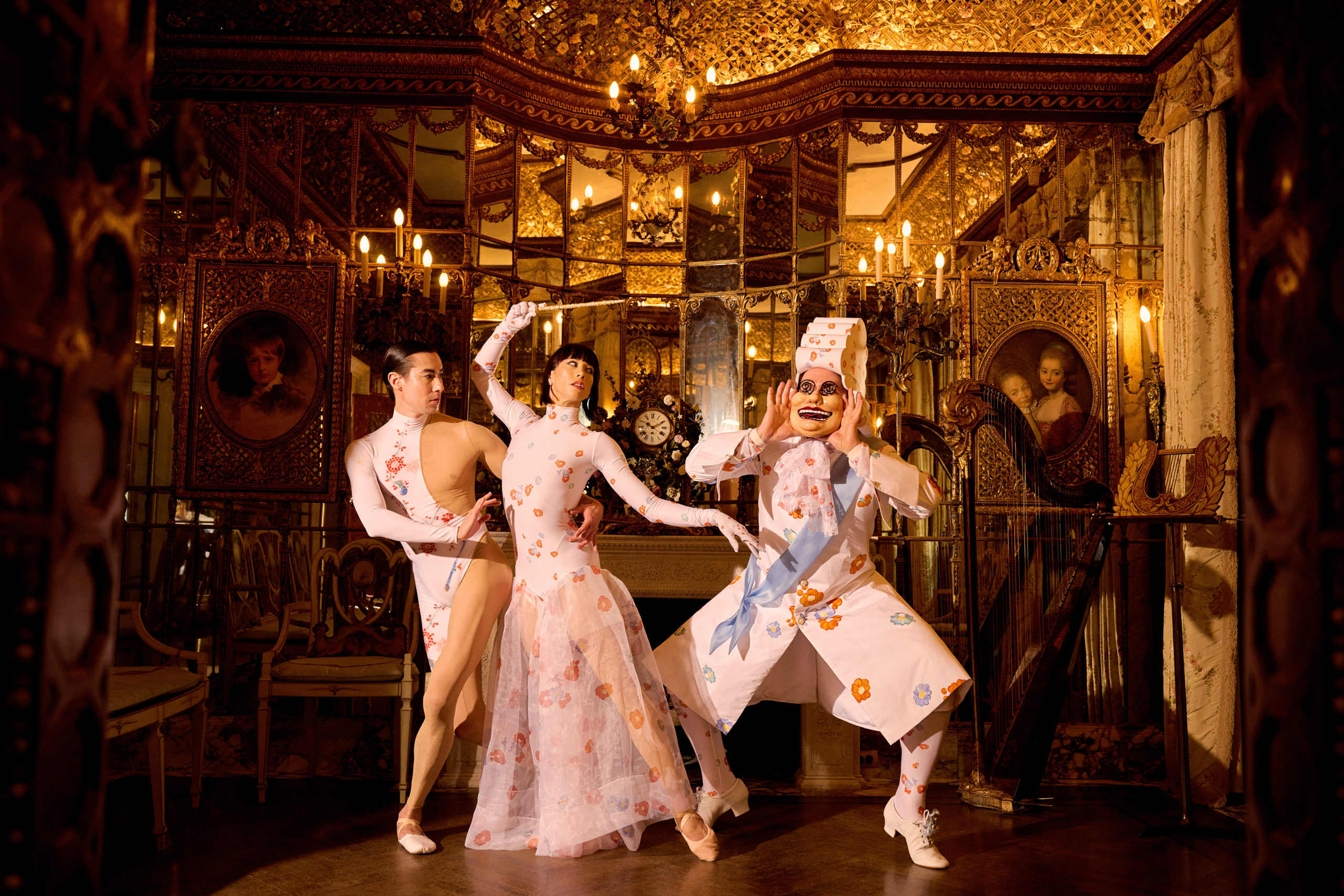 In an ornate, gilded room full of mirrors, Daniel Appelbaum, Georgina Pazcoguin and Tyler Haynes pose in floral-patterned costumes for Ballet des Porcelaines. Hanes wears a mask with a large, exaggerated smile and a tall hat.