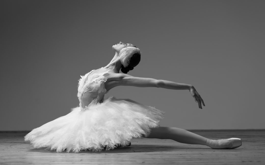 Olga Smirnova, wearing a white Swan Lake costume tutu and feathered headpiece, sits on her right knee and extends her left leg straight behind her on the floor. She bends her upper body back deeply, reaching her arms back and looking up towards the ceiling.