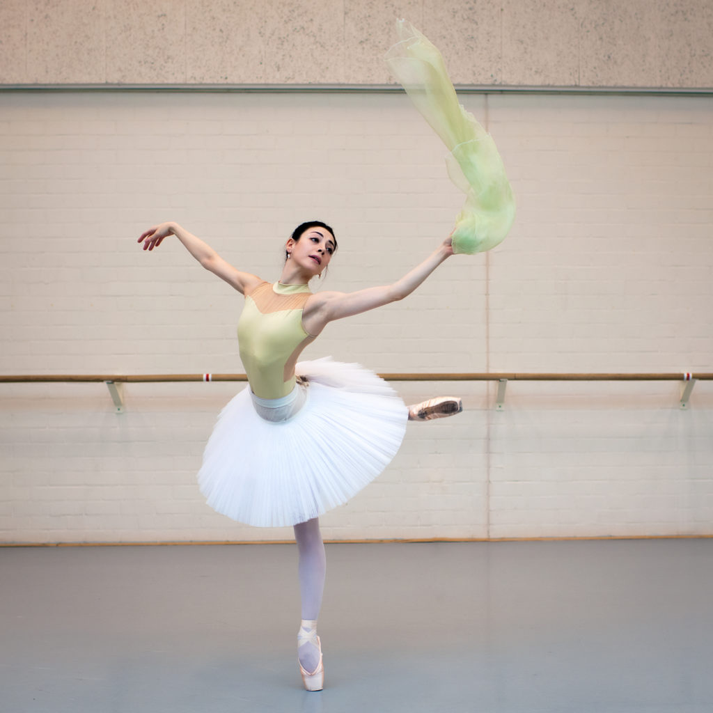 Maia Makhateli wears a light green leotard, white practice tutu and pink tights and pointe shoes. She practices an attitude derriere on pointe with her right leg behind her and waves a light green scarf with her left hand.