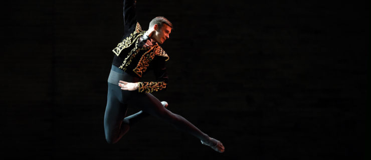 Oleksii Potiomkin, wearing black tights and a black and gold bolero-style jacket, jumps high into the air with his right leg in passé, arching his body over the the left. He holds his right arm up above his head and holds his waist with his left hand.