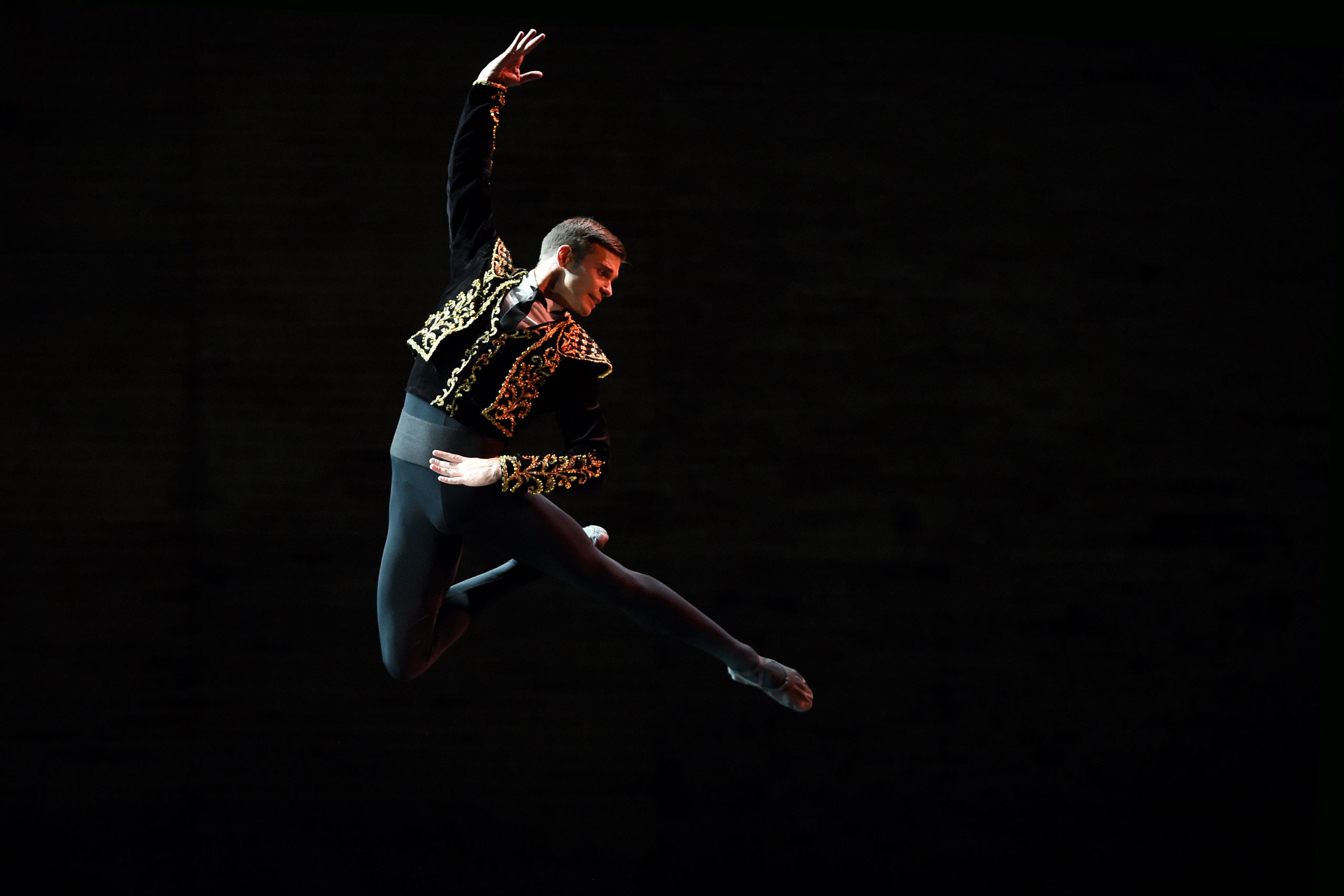 Oleksii Potiomkin, wearing black tights and a black and gold bolero-style jacket, jumps high into the air with his right leg in passé, arching his body over the the left. He holds his right arm up above his head and holds his waist with his left hand.