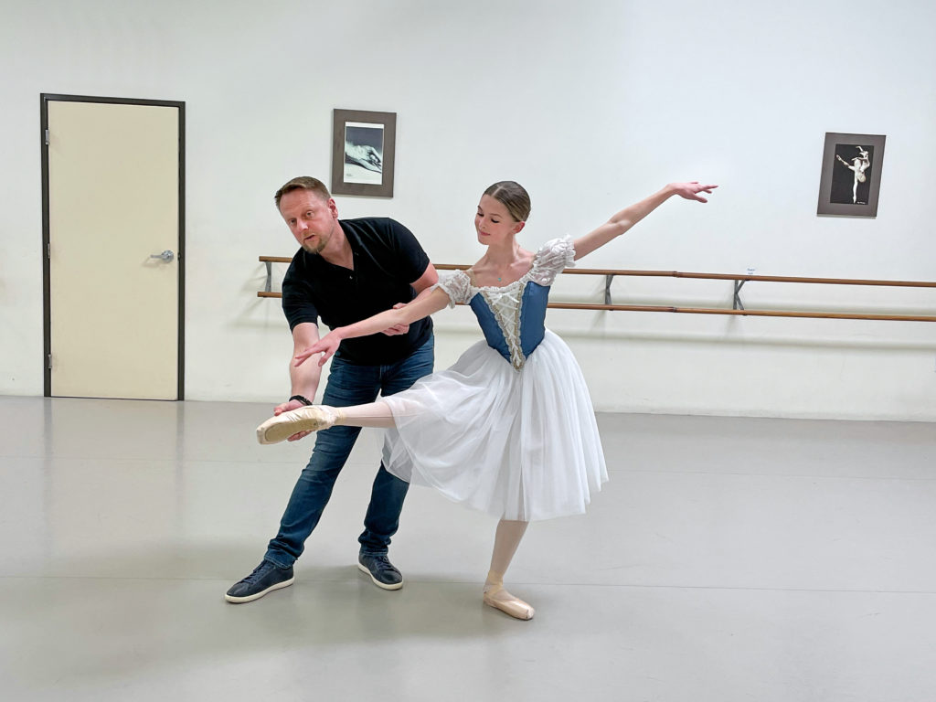 Dmitri Kulev stands behind his student, Natalie Steele, and adjusts her lifted right foot as she poses in a knne-height battement devant in effacé. She extends her right arm in line with her right leg and reaches her left arm behind her to create a diagonal. She wears a peasant dress costume with a white knee-length tutu and blue bodice; Kulev wears jeans, a black polo shirt and sneakers.