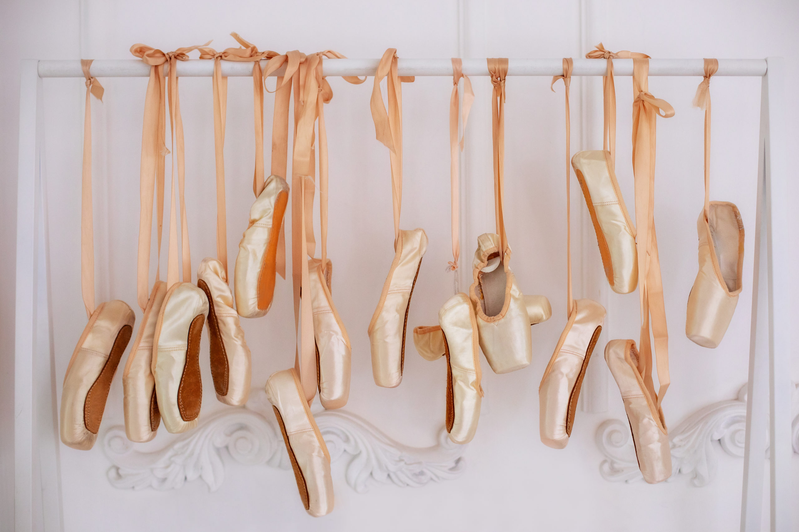 New pointe shoes with satin ribbons hang on a white metal barre.
