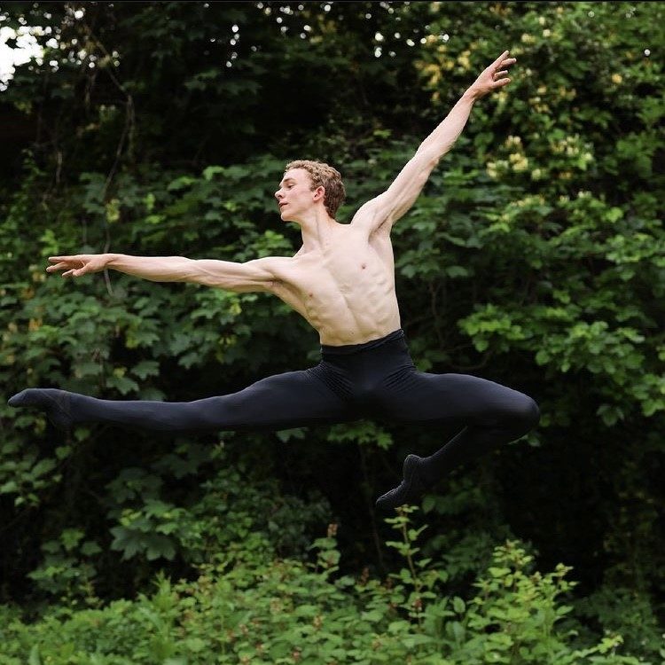 Henry Griffin, a muscular young man wearing black tights, ballet shoes and no shirt, leaps outside in a green yard.