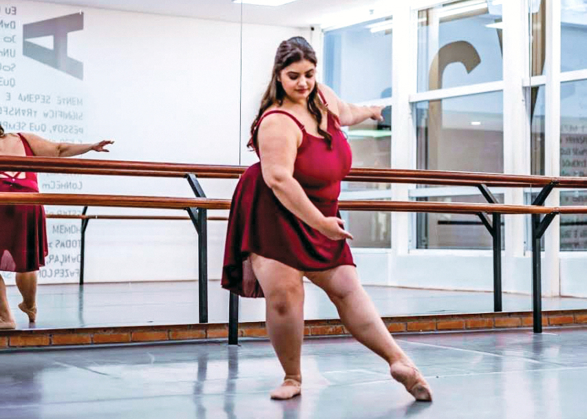 Júlia Del Blanco wears a burgundy camisole leotard and matching ballet skirt and pink ballet slippers. She is shown in a dance studio doing a tendu devant in effacé with her left leg extended and her right leg in plié.